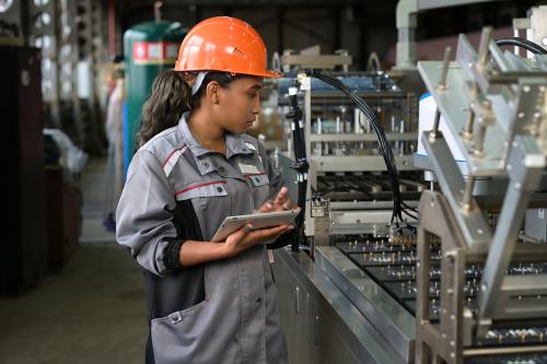 A young black manufacturing worker controls an assembly line in a factory, making notes on a tablet. Young woman in hard hat watches the work process